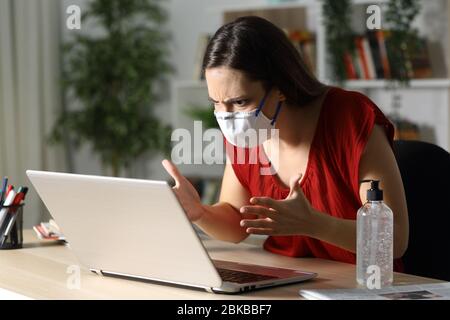 Une fille en colère vérifiant un ordinateur portable sur un confinement du coronavirus assis sur un bureau à la maison dans la nuit Banque D'Images