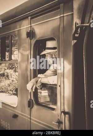 Sépia près de l'homme des années 1940 dans le chapeau penchant et regardant hors du train à vapeur britannique vintage en quittant la gare, chemin de fer Severn Valley des années 1940 guerre événement d'été. Banque D'Images