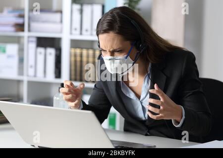 Une femme en colère et démarchée qui regarde un ordinateur portable évitant le covid-19 avec un masque assis sur un bureau au bureau Banque D'Images