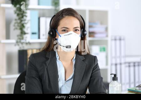 Une femme de télévendeur heureuse qui pose sur l'appareil photo en évitant le covid-19 avec masque assis au bureau Banque D'Images