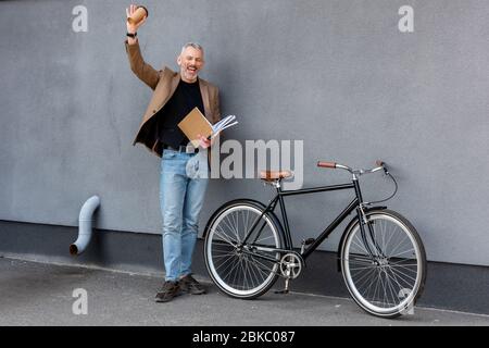 un homme d'affaires heureux se révechant la main tout en tenant la tasse et le dossier en papier et en se tenant près de la bicyclette Banque D'Images