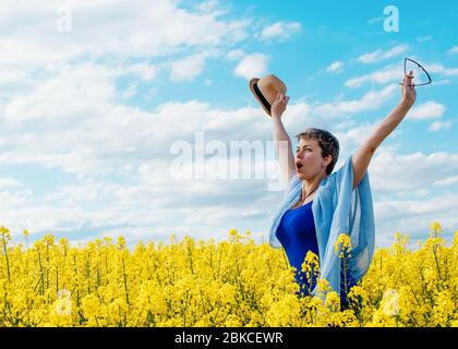 Une femme heureuse portant un haut bleu et un Jean lui lève les mains élevé contre le ciel bleu et les champs de colza jaune Banque D'Images