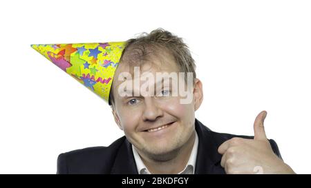 Portrait de jeune homme endormi avec un hangover donnant des pouces vers le haut, sourires. Émotions. Un homme d'affaires en costume et un bonnet festif à la porte de l'appareil photo. Fond blanc. Fête d'anniversaire de bureau Banque D'Images