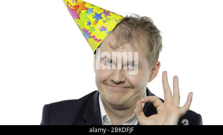 Portrait de l'homme jeune et endormi de la planche avec un hangover montrant le signe OK et le sourire. Émotions. Un homme d'affaires en costume et un bonnet festif à la porte de l'appareil photo. Fond blanc. Concept de fête de bureau Banque D'Images