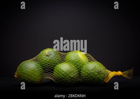 Fruits à la chaux vert et jaune dans un sac à filet contrasté sur un fond gris foncé. Studio produit dynamique faible durée de vie. Banque D'Images