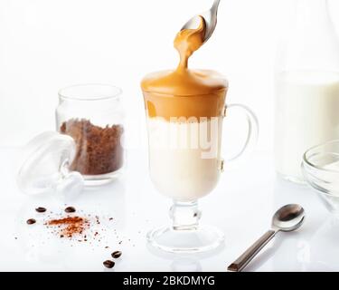 Café Dalgona maison tendance dans une tasse en verre avec des ingrédients, café instantané, lait et suger isolés sur fond blanc. Recette populaire de drin coréen Banque D'Images