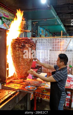 Un serveur coupe des portions de viande d'un grand shawarma qui cuisine sur un feu dans un restaurant de rue dans le marché municipal de Merida. Banque D'Images