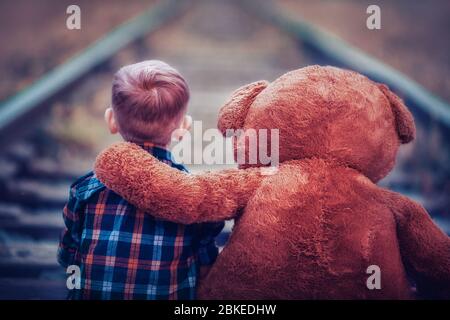 Un petit garçon avec un grand ours en peluche est assis sur les voies ferrées. Solitude et tristesse d'un enfant. Le problème des enfants abandonnés. Banque D'Images