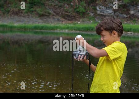 garçon de 9 ans debout dans l'eau, lave l'appareil photo numérique. Enfance, puériculture, mauvais appareil photo de nettoyage. Mesures préventives de propagation des virus. Banque D'Images