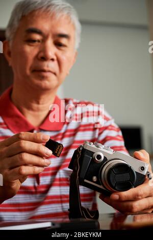 Homme asiatique mature qui prend une carte mémoire hors de l'appareil photo pour télécharger des photos sur l'ordinateur Banque D'Images