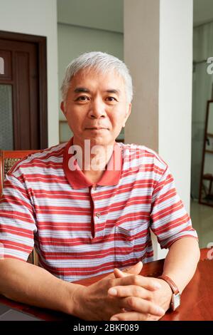 Positif homme chinois âgé assis au bureau en bois à la maison et regardant la caméra Banque D'Images