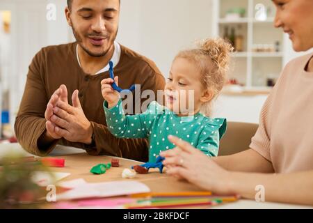 Petite fille portant une robe à pois poka assise à table avec ses parents, en faisant des modèles simples à l'aide de pâte à jouter Banque D'Images