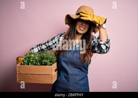 Jeune femme agriculteur brunette portant un tablier et une boîte de maintien de chapeau avec des plantes stressées avec la main sur la tête, choquée avec la honte et le visage surprise, Banque D'Images