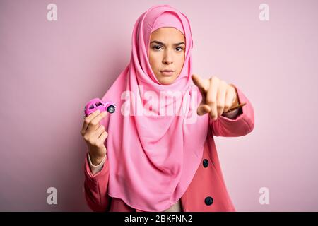 Jeune fille portant le hijab musulman tenant un petit jouet de voiture sur un fond rose isolé pointant avec le doigt à l'appareil photo et à vous, signe de main, positif Banque D'Images