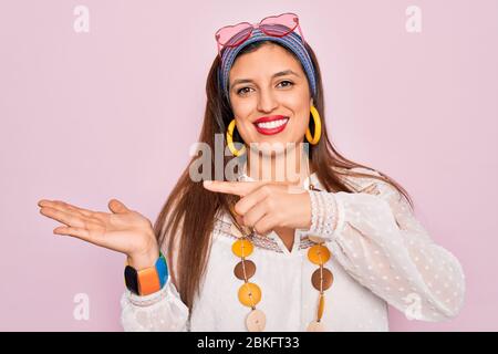 Jeune femme hippie hispanique portant un style boho de mode et des lunettes de soleil sur fond rose stupéfait et souriant à l'appareil photo tout en présentant avec la main Banque D'Images