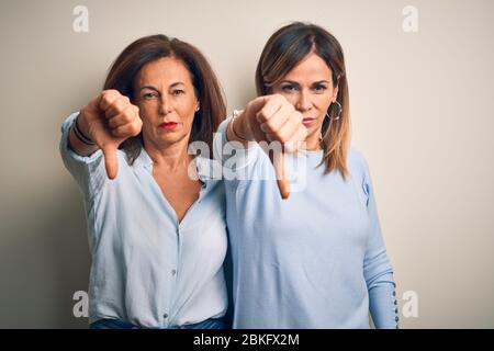 Beau couple de sœurs d'âge moyen se tenant sur un fond blanc isolé, mécontent et fâché montrant le rejet et négatif avec les pouces le font Banque D'Images