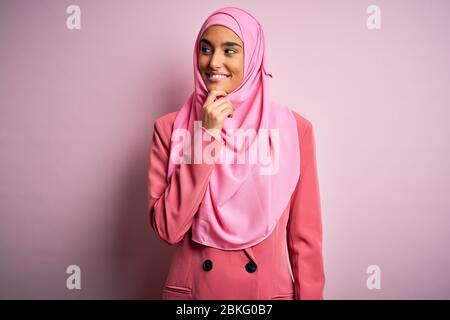 Jeune femme d'affaires brunette, vêtue de hijab musulman rose et de veste d'affaires avec main sur le menton, pensant à la question, expression pensive. SM Banque D'Images