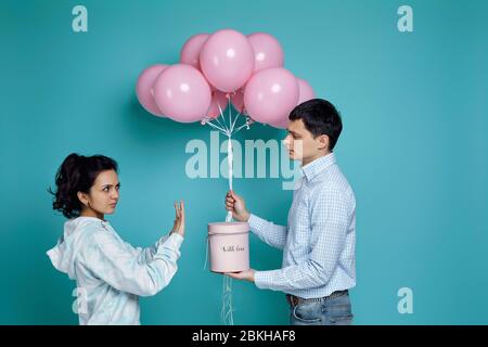 jeune homme présentant boîte cadeau et ballons d'air rose à sa fille bouleversée frite sur fond bleu. la femme refuse un cadeau Banque D'Images