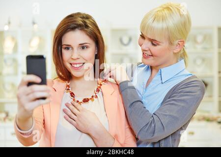 Une femme souriante au bijoutier fait selfie avec smartphone tout en essayant sur un collier Banque D'Images