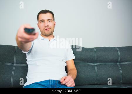 Un jeune homme s'assoit dans la chambre pendant la quarantaine. Un gars qui utilise la télécommande tient la télécommande dans une main. Guy s'assoir seul et changer de chaîne de télévision. Banque D'Images