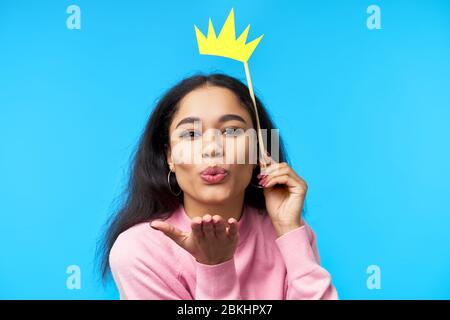 Jolie femme noire envoie un baiser d'air avec une couronne en papier sur un bâton sur fond bleu. Partie, émotions concept Banque D'Images