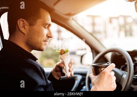Arrêt pour un morceau à manger . L'homme mange un en-cas dans la voiture et boit du café ou du thé. Banque D'Images