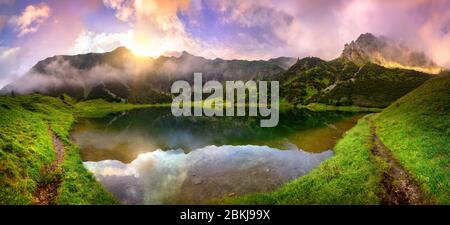 Magnifique lever de soleil sur un lac dans les Alpes avec des montagnes, des nuages colorés et des wafts de brume reflétés dans l'eau claire et les chemins menant à travers le Banque D'Images
