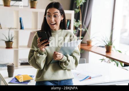 Image d'une belle jolie jeune fille excitée choquée étudiant à l'intérieur tenant le presse-papiers à l'aide du téléphone portable. Banque D'Images