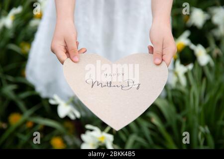 Joyeux fête des mères écrit sur une carte en forme de coeur tenue par une fille sur fond de fleurs et d'herbe Banque D'Images