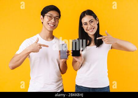 Portrait d'un jeune couple asiatique très joyeux portant des vêtements décontractés isolés sur fond jaune, montrant un téléphone portable à écran vide Banque D'Images