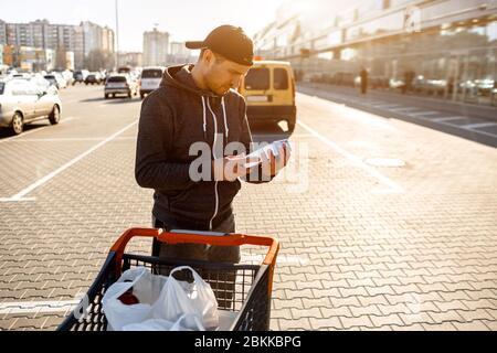 Un homme lit la composition des ingrédients alimentaires sur l'emballage dans le parking d'un centre commercial ou d'un supermarché. Près de elle se trouve une poussette pour Banque D'Images