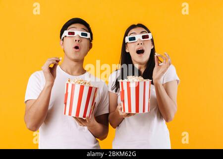 Image d'un couple multinational choqué dans des lunettes en relief mangeant du pop-corn tout en regardant un film isolé sur fond jaune Banque D'Images