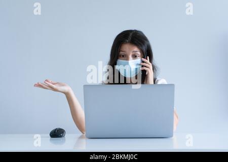 La fille travaille de la maison pendant la quarantaine. Pandémie du virus Corona. Une fille dans un masque chirurgical travaille sur un ordinateur portable et boit du café. Travail à distance. Banque D'Images