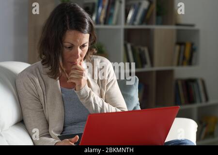 Femme sérieuse concentrée d'âge moyen regardant du contenu vidéo en ligne sur un ordinateur portable assis sur le canapé la nuit à la maison Banque D'Images