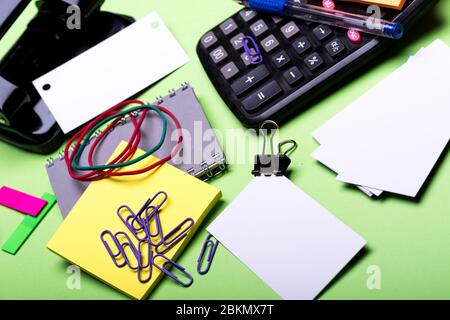 Calculatrice et papeterie. Outils Office sur fond vert, gros plan. Carnet de couleur grise, perforateur, cartes de visite, notes adhésives, élastiques et clips. Concept de travail professionnel et de bureau. Banque D'Images