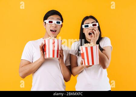 Image d'un couple multinational effrayé dans des lunettes en relief mangeant du pop-corn tout en regardant un film isolé sur fond jaune Banque D'Images