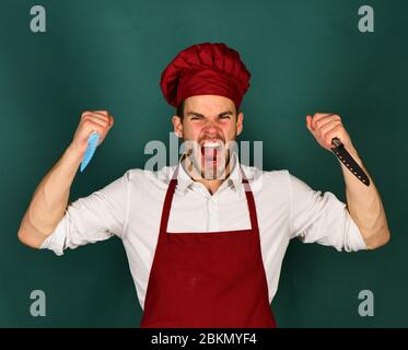 Homme dans le chapeau de cuisinier et le tablier fouets les couteaux. Le chef au visage fou tient des couteaux bleus et métalliques sur fond vert foncé. Ustensiles de cuisine et concept de cuisine. Cuisine Banque D'Images