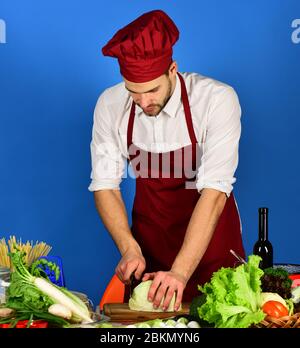 Ustensiles de cuisine et concept de cuisine. Chef avec chops de visage très occupés chou avec couteau sur fond bleu. Cuisiner travaille dans la cuisine près de la table avec des légumes et des outils. Homme dans le chapeau de cuisinier et le tablier coupe le chou Banque D'Images