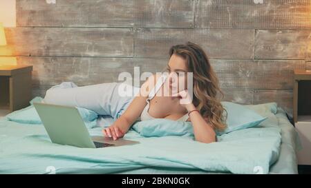 Zoom arrière de la belle femme portant un pyjama travaillant sur un ordinateur portable avant le coucher. Chien heureux. Banque D'Images