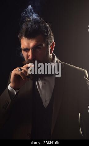 Un homme avec une barbe tient le cigare sur fond gris. Un homme d'affaires au visage confiant fume le cigare cubain. Concept d'affaires et de mauvaise habitude. Guy en costume classique a cinq minutes de pause pour fumer. Banque D'Images