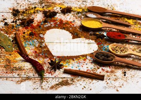 Cuillères à bois avec poivre, paprika, curcuma, bâtons de cannelle, étoile anis et assaisonnement épaté. Composition du condiment faisant la forme du coeur. Ensemble d'épices sur fond de bois blanc. Concept de l'art alimentaire Banque D'Images