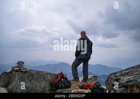 Jeune randonneur indien chauve, vêque de vêtements chauds, debout au bord d'un rocher de montagne et profitant de la vue imprenable Banque D'Images