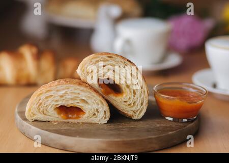 Deux moitiés de croissant fraîchement poêlé sur bois avec confiture d'abricot maison délirante. Fond de cappuccino. Petit déjeuner Banque D'Images