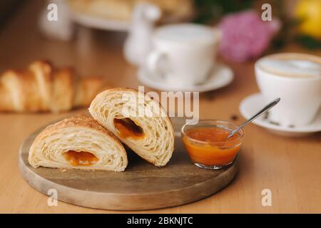 Deux moitiés de croissant fraîchement poêlé sur bois avec confiture d'abricot maison délirante. Fond de cappuccino. Petit déjeuner Banque D'Images