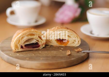 Deux moitiés de croissant fraîchement poêlé sur une planche de bois avec une confiture différente. Fond de cappuccino Banque D'Images