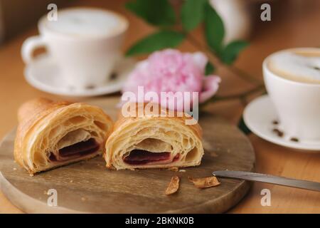 Deux moitiés de croissant fraîchement poêlé sur bois avec confiture de framboise maison délirante. Fond de cappuccino. Pivoine rose Banque D'Images