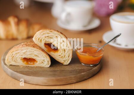Deux moitiés de croissant fraîchement poêlé sur bois avec confiture d'abricot maison délirante. Fond de cappuccino Banque D'Images