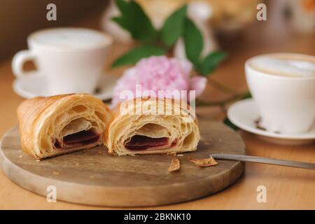Deux moitiés de croissant fraîchement poêlé sur bois avec confiture de framboise maison délirante. Fond de cappuccino. Pivoine rose Banque D'Images
