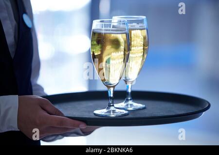 Les mains d'un serveur vêtu d'une veste noire et d'une chemise blanche tenant deux verres de champagne sur un plateau Banque D'Images