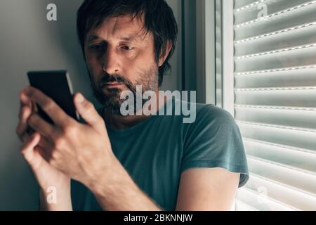 Homme messagerie texte sur téléphone mobile dans l'intimité de sa chambre à côté de la fenêtre avec volets fermés, foyer sélectif Banque D'Images